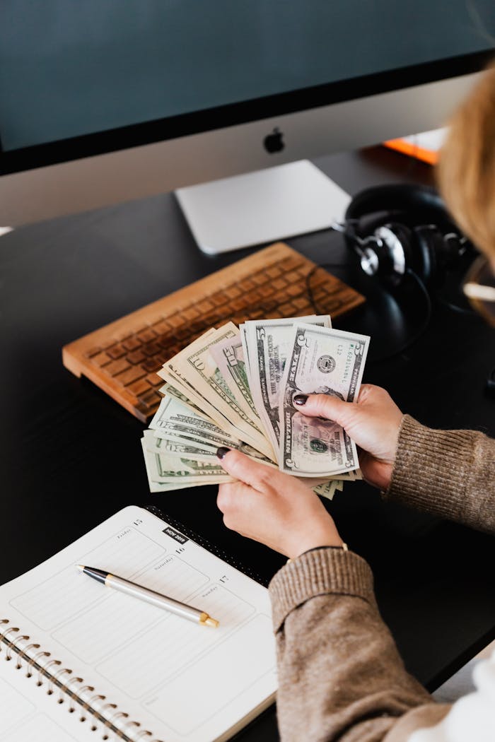 vision Close-up of hands counting dollar bills at a desk with computer and planner.