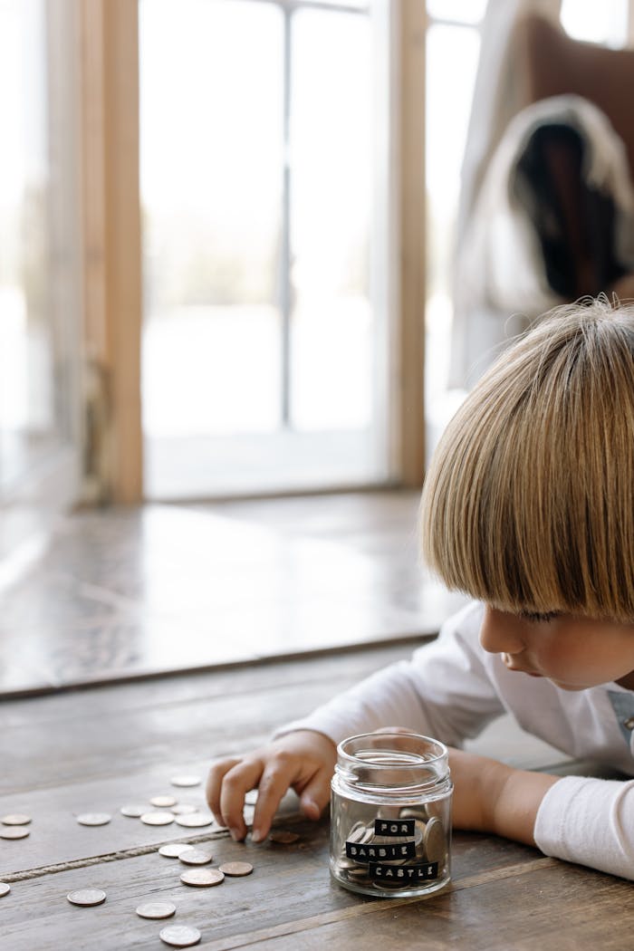 services-03 A young boy counting coins and placing them into a jar labeled 'for barbie castle' indoors on wooden flooring.